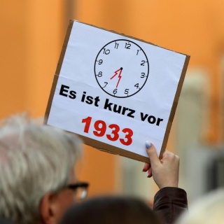 Ein Demonstrant hält ein Schild in die Höhe, auf dem ein Zifferblatt einer Uhr zu sehen ist und "Es ist kurz vor 1933" steht.
