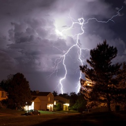 Über einem Wohngebiet steht eine Gewitterwolke, aus der ein weit verzweigter Blitz hervorschlägt.