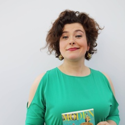 Nora Gomringer in einem türkis-grünen Kleid vor grauem Hintergrund. Sie hält ein Buch in der Hand und grinst in die Kamera.
